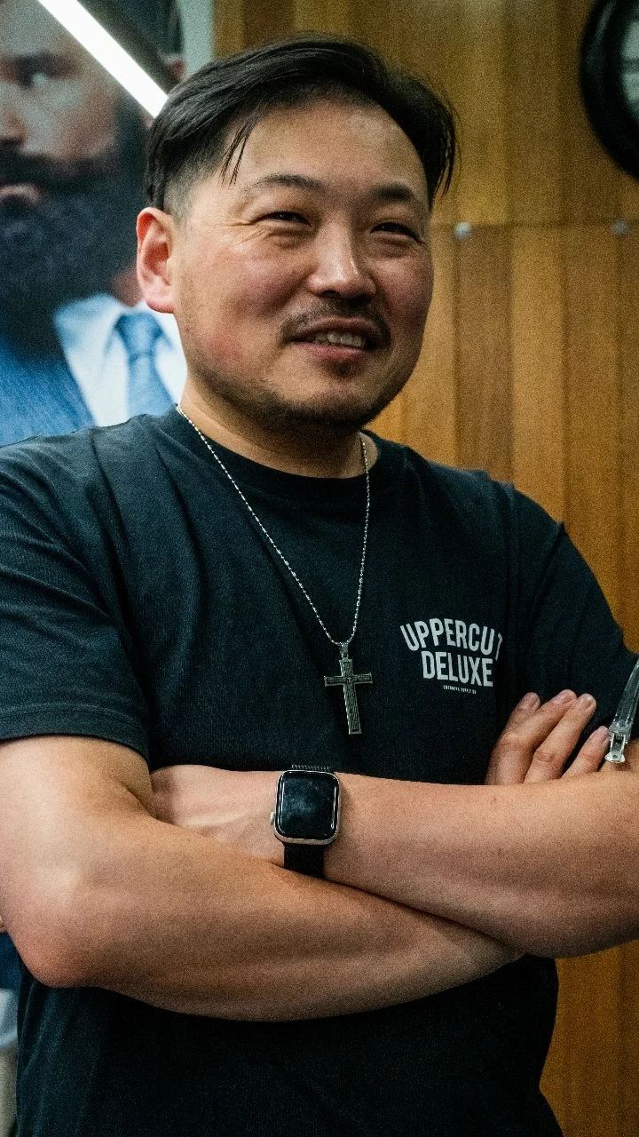 Jay, owner of HO2 Barber Shop, arms crossed and smiling in front of the timber wall in his shop